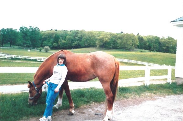 A white girl wearing a sweatshirt and breeches with short dark hair leaning against a chestnut mare with three white legs, grazing on a bit of grass by an outdoor ring. 